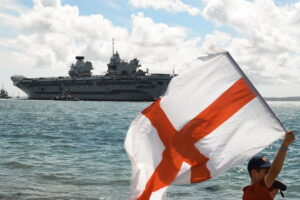Το αεροπλανοφόρο HMS Prince of Wales κατά την αναχώρησή του από το Πόρτσμουθ το 2025, με τη σημαία της Αγγλίας στο προσκήνιο.