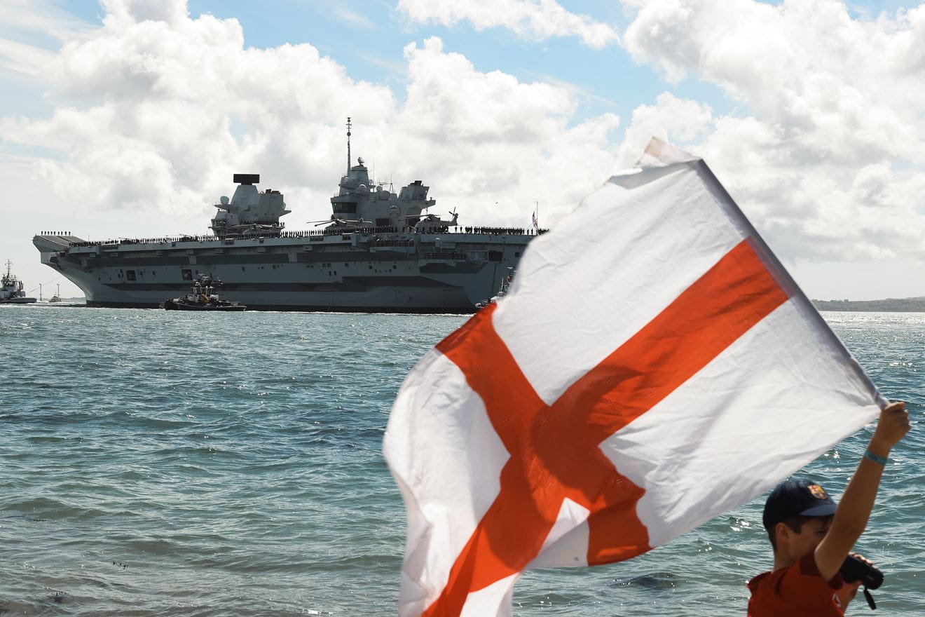 Το αεροπλανοφόρο HMS Prince of Wales κατά την αναχώρησή του από το Πόρτσμουθ το 2025, με τη σημαία της Αγγλίας στο προσκήνιο.